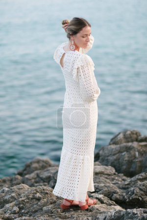 Téléchargez les photos : Vue latérale d'une jeune touriste tranquille en robe blanche romantique touchant les cheveux et regardant vers le bas tout en se tenant sur le bord de mer rocheux pendant les vacances d'été - en image libre de droit