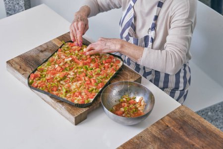 Foto de Desde arriba de la cosecha anónimo jefe femenino en delantal cocinar pizza en la tabla de cortar mientras está sentado en la mesa con un tazón lleno de verduras frescas cortadas - Imagen libre de derechos