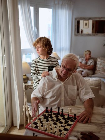 Téléchargez les photos : Garçon pensif debout près de grand-père concentré jouant jeu d'échecs intéressant à la table dans le salon léger avec femme à la maison - en image libre de droit
