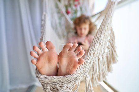 Photo for Adorable boy resting in wicker hammock on balcony while surfing internet via smartphone during warm sunny day - Royalty Free Image