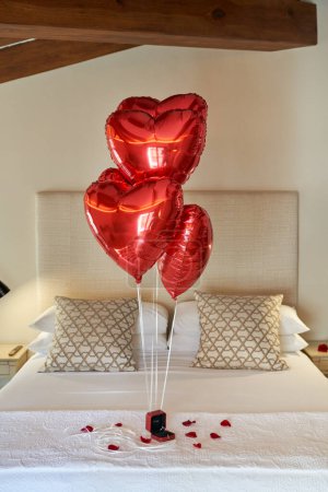 Photo for Hotel bedroom decorated with red heart shaped balloons and scattered petals of roses and gift box with engagement ring placed on comfortable bed for romantic evening of loving couple - Royalty Free Image