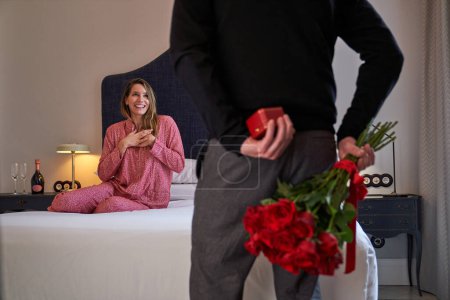 Téléchargez les photos : Dame positive en vêtements de nuit sur le lit en regardant le petit ami de la culture cachant boîte cadeau et roses rouges - en image libre de droit