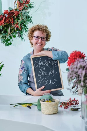 Photo for Professional female florist with curly hair smiling and looking at camera while standing at white table with flowers and demonstrating board with inscription during workday in modern floral shop - Royalty Free Image