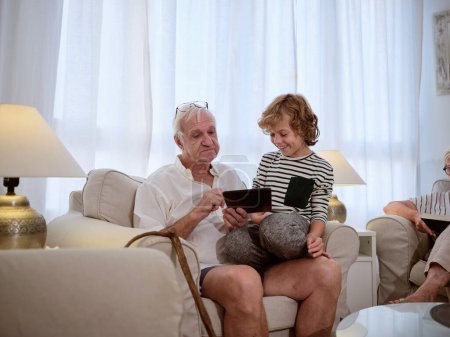 Téléchargez les photos : Homme âgé surfant tablette moderne avec petit-fils positif tout en étant assis ensemble sur un canapé confortable dans le salon léger à la maison - en image libre de droit