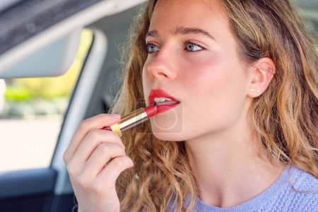 Photo for Sensitive female driver with blue eyes and wavy hair applying red lipstick in car on sunny day - Royalty Free Image