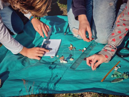 Téléchargez les photos : De dessus des enfants de préadolescence de culture mesurant des morceaux ramassés assortis de déchets de plastique à l'aide d'un étrier numérique et d'une règle placés sur un tissu bleu sur le sol - en image libre de droit