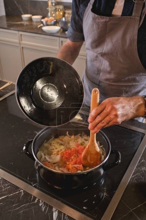 Téléchargez les photos : Crop man en tablier gris debout à la cuisinière et mélanger la sauce tomate avec des nouilles tout en cuisinant dans la cuisine moderne à la maison - en image libre de droit