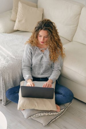 Photo for From above of young woman with curly hair in casual clothes sitting on pillow on floor and browsing laptop - Royalty Free Image