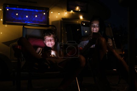 Téléchargez les photos : Gai femme et garçon souriant et naviguant sur les téléphones cellulaires tout en se refroidissant sur des chaises longues camping-car extérieur à la nuit noire - en image libre de droit