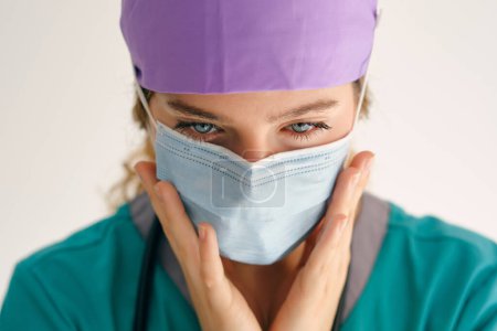Photo for Professional female doctor in medical scrubs wearing protective mask and touching cheeks on beige background - Royalty Free Image