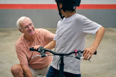 Téléchargez les photos : Homme âgé positif en tenue décontractée et lunettes avec garçon anonyme en casque sur vélo passer du temps ensemble sur le parking - en image libre de droit