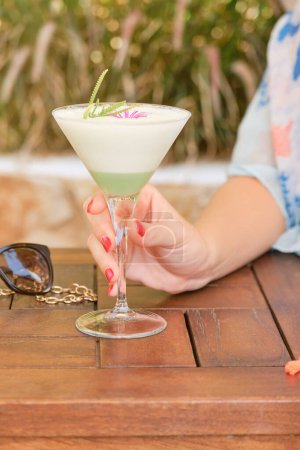 Téléchargez les photos : Crop dame anonyme avec verre de cocktail frais sur table en bois près de plantes vertes floues - en image libre de droit