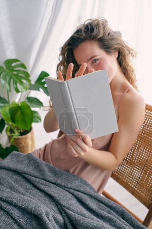 Téléchargez les photos : D'en haut de la jeune femme aux cheveux longs assis sur une chaise en osier sous une couverture chaude et lisant un livre intéressant près de la fenêtre et de la plante de Monstera à la maison - en image libre de droit