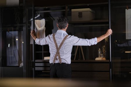 Photo for Back view of man in trendy outfit with suspenders opening glass walls of wardrobe in dim light - Royalty Free Image