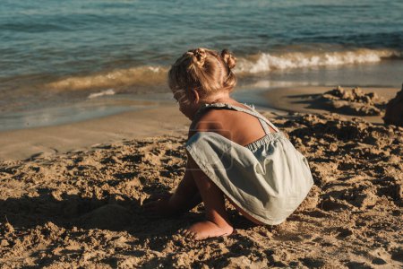 Téléchargez les photos : Petite fille en vêtements décontractés jouant avec le sable près de la mer ondulante sur la plage pendant la journée ensoleillée - en image libre de droit