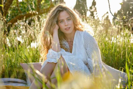 Foto de Atractiva joven rubia en la espalda iluminada usando elegante vestido blanco tocando el pelo ondulado y mirando a la cámara mientras descansa en el césped verde herboso en el soleado jardín de verano - Imagen libre de derechos