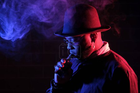 Photo for Side view of bearded man in hat smoking vape while standing with sunglasses in dark room - Royalty Free Image