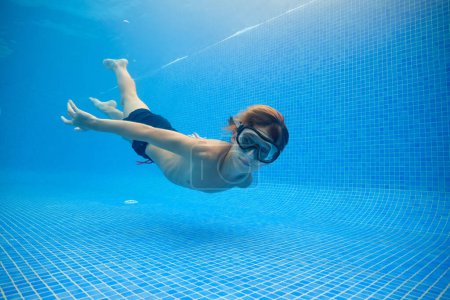 Foto de Vista lateral de cuerpo completo del niño en gafas nadando en la piscina azul limpia cerca de la parte inferior y mirando a la cámara - Imagen libre de derechos