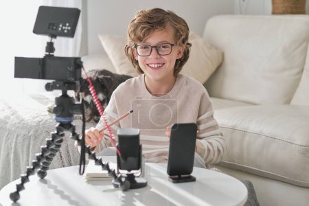 Téléchargez les photos : Enfant souriant en vêtements décontractés assis à la table avec un équipement professionnel en cours de préparation pour l'enregistrement vidéo pour le blog dans le salon moderne à la maison - en image libre de droit