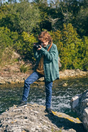 Téléchargez les photos : Vue latérale complète du corps d'un enfant concentré prenant des photos avec un appareil photo professionnel tout en se tenant debout sur la pierre près d'un ruisseau coulant - en image libre de droit