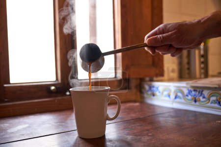 Photo for Crop anonymous man pouring fresh brewed aromatic coffee from cezve into white ceramic cup placed on wooden table near window - Royalty Free Image