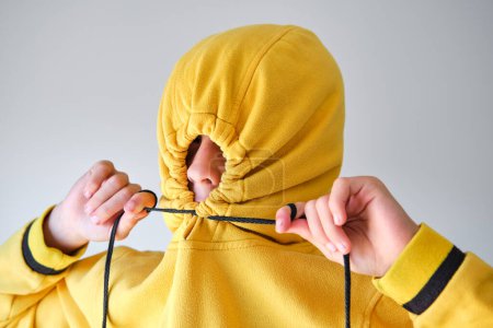 Foto de Niño con capucha amarilla ajustando los cordones en la capucha sobre la cabeza revelando la nariz contra el fondo gris - Imagen libre de derechos