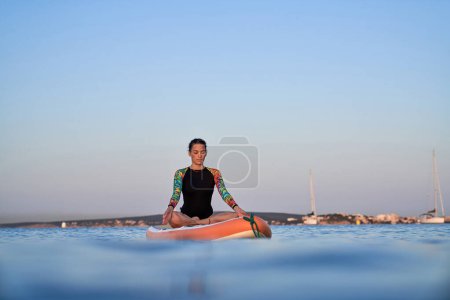 Photo for Full body of young Hispanic woman in swimwear meditating in lotus pose while practicing yoga on SUP board in calm seawater under cloudless evening sky - Royalty Free Image