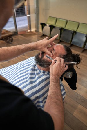 Photo for High angle of crop anonymous barber using straight blade to cut beard of male client while working in barbershop - Royalty Free Image