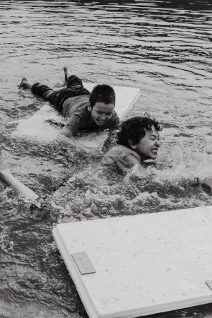 Téléchargez les photos : Noir et blanc de garçons gais allongés sur une planche plate tout en nageant sur une piscine ondulée rugueuse en plein jour - en image libre de droit