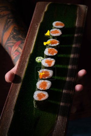 Téléchargez les photos : De dessus du serveur tatoué anonyme de récolte servant ensemble de délicieux rouleaux d'uramaki avec du saumon sur un plateau en bois - en image libre de droit