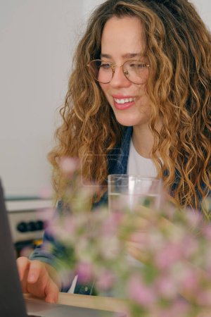 Foto de Joven freelancer en gafas sentada en la mesa con portátil y mirando a la pantalla mientras trabaja en línea en el proyecto - Imagen libre de derechos