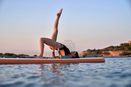 Téléchargez les photos : Corps complet de la jeune athlète féminine en maillot de bain prenant Setu Bandha Sarvangasana pose avec la jambe levée tout en pratiquant le yoga sur le conseil SUP dans l'océan ondulation au coucher du soleil - en image libre de droit
