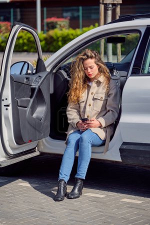 Photo for Full length beautiful female in jeans and coat using mobile phone while sitting in car with opened door on sunny day - Royalty Free Image