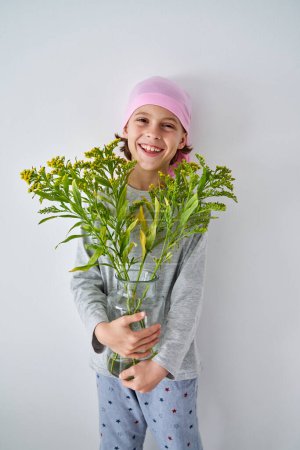 Photo for Glad kid in casual clothes and pink headband holding Solidago and looking at camera while standing near white wall - Royalty Free Image