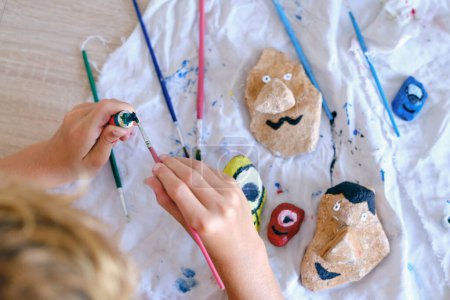 Photo for From above of crop faceless kid using watercolors while painting funny faces on stones at table at home - Royalty Free Image