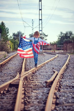 Téléchargez les photos : Enfant tenant drapeau des États-Unis avec le sourire et marchant le long du chemin de fer près de commutateur sous un ciel bleu nuageux - en image libre de droit