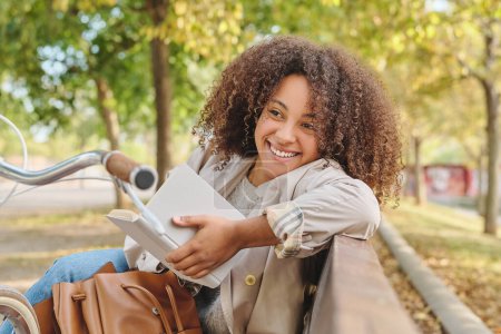 Téléchargez les photos : Une jeune femme hispanique positive avec une coiffure afro assise sur un banc dans un parc près d'un vélo et lisant un livre tout en regardant ailleurs - en image libre de droit