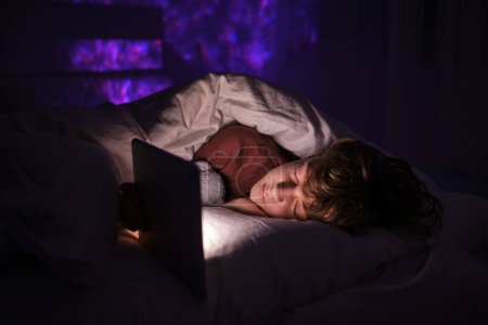 Foto de Niño tranquilo con los ojos cerrados durmiendo cerca de la tableta brillante mientras está acostado debajo de la manta en la cama en el dormitorio con poca luz al final de la noche - Imagen libre de derechos