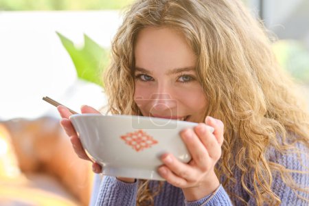 Téléchargez les photos : Jeune femme blonde heureuse mangeant des ramen du bol et regardant la caméra sur fond flou - en image libre de droit