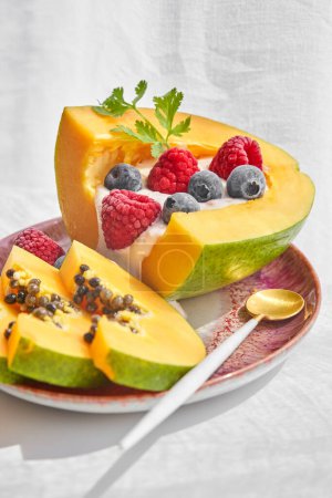 Téléchargez les photos : Portion de papaye délicieuse servie sur une assiette en céramique avec cuillère et décorée de yaourts framboises et bleuets sur fond blanc - en image libre de droit
