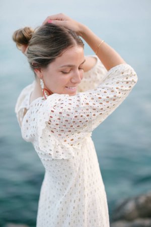 Téléchargez les photos : Positif jeune femme féminine en robe blanche élégante souriant et regardant vers le bas tout en touchant la tête sur le bord de mer rocheux au coucher du soleil - en image libre de droit