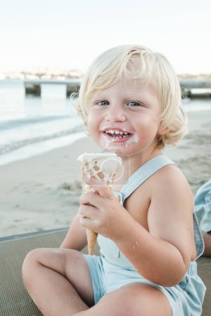 Photo for Adorable little boy in casual clothes smiling and looking at camera while sitting on beach and eating ice cream - Royalty Free Image
