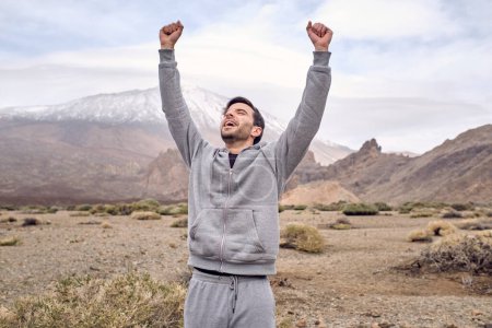 Photo for Cheerful male traveler raising arms while celebrating achievement of climbing up on mountain during journey in Tenerife in Canary Islands - Royalty Free Image