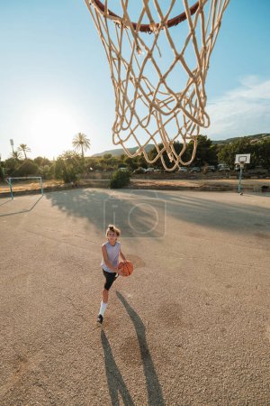 Foto de Desde arriba de chico activo en ropa deportiva jugando baloncesto en el patio de recreo contra el cielo en el día soleado - Imagen libre de derechos