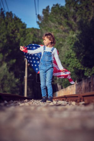 Téléchargez les photos : Toute la longueur de l'enfant regarde loin avec intérêt tout en célébrant la fête de l'indépendance des États-Unis sur le chemin de fer contre les bois verts au soleil - en image libre de droit