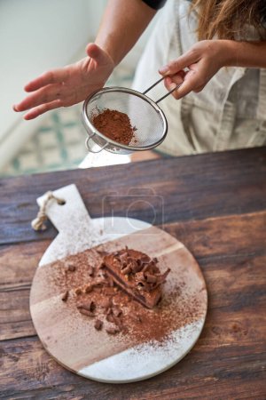 Foto de De arriba anónimo hembra tamizado cacao en polvo en sabroso pastel de chocolate con colador a bordo en la mesa de madera - Imagen libre de derechos