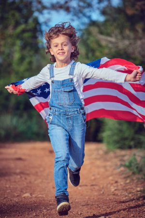Foto de Cuerpo completo de adorable chico de contenido con bandera americana mirando a la cámara mientras corre por la carretera en el campo de verano - Imagen libre de derechos