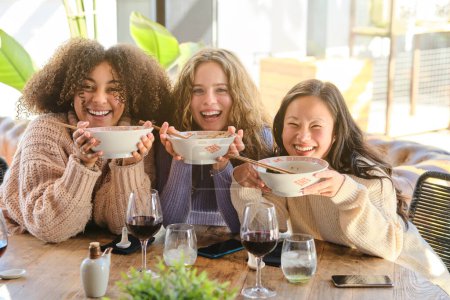 Foto de Mujeres multirraciales positivas mirando a la cámara mientras se sientan en la mesa con cuencos de ramen y beber vino - Imagen libre de derechos
