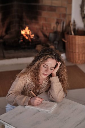 Téléchargez les photos : Femme inspirée en vêtements décontractés assis à une table en bois près d'une cheminée en feu et prenant des notes dans un bloc-notes dans un salon confortable à la maison - en image libre de droit