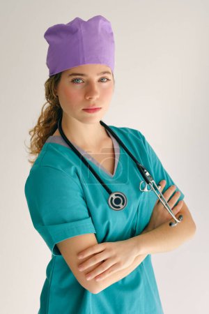 Photo for Young woman in medical uniform and purple cap with stethoscope on neck looking at camera with crossed arms in studio - Royalty Free Image